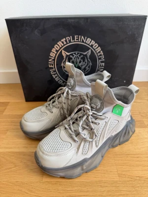 Grå sneakers från Plein Sport - Chunky sneakers i grått från Plein Sport med mesh och syntetdetaljer, tjock sula i mörkgrått och coola loggor på plös och häl. Skorna har snörning, dragflikar och gröna detaljer. Perfekta för dig som gillar streetwear och statement-skor. Använd ett par gånger, mycket gott skick, medföljer box. Äkta 