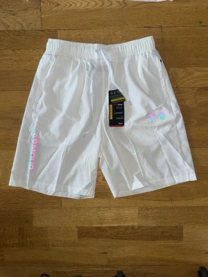 Vita shorts från Under Armour med reflex logga - Säljer ett par vita shorts från Under Armour med elastisk midja och snörning. Shortsen har holografiska detaljer i rosa, grön och orange samt logga och text på ena benet. Perfekta för sommarens alla aktiviteter. Det står storlek M men det passar både M och S.
