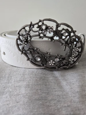 Vitt bälte med dekorativt spänne - Snyggt vitt bälte med präglat mönster och ett stort, antik-inspirerat spänne i metall med blommiga detaljer och glittrande stenar. Perfekt accessoar för att lyfta din outfit med en unik touch.