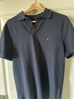 Mörkblå pikétröja från Tommy Hilfiger - Klassisk mörkblå pikétröja från Tommy Hilfiger med korta ärmar och liten logga på bröstet. Tröjan har krage och knappar vid halsen, perfekt för en clean och stilren look.