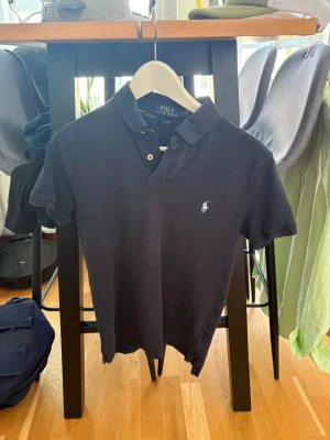 Svart pikétröja från Ralph Lauren - Klassisk svart pikétröja från Ralph Lauren med vit broderad logga på bröstet. Tröjan har korta ärmar, krage och knappar vid halsen. Perfekt för en stilren och avslappnad look.
