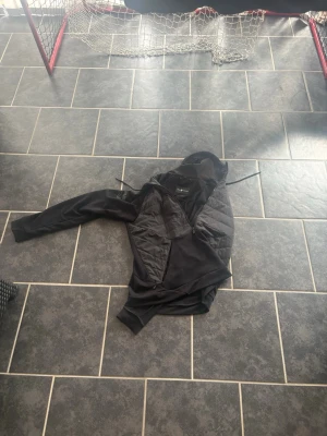 Svart hybridjacka med huva - Svart hybridjacka med quiltade partier på sidorna och mjuka ärmar. Jackan har huva med dragsko och dragkedja framtill. Perfekt för lager-på-lager och streetwear-stil.