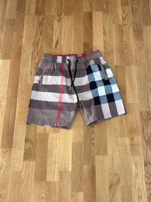 Rutiga badbyxor från Burberry - Säljer ett par snygga badbyxor från Burberry med klassiskt rutmönster i beige, svart, vitt, ljusblått och en röd detalj. De har elastisk midja med dragsko och sidofickor. Perfekta för stranden eller poolen.