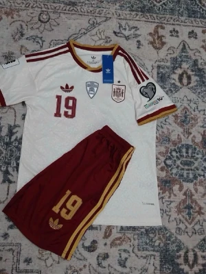Spanien matchställ Adidas nr 19 - Säljer ett officiellt matchställ från Spaniens landslag med nummer 19 och Lamine Yamal på ryggen. Tröjan är vit med vinröda detaljer och guldkanter, och shortsen är vinröda med gula ränder. Tillverkad i lätt och ventilerande material, perfekt för fotbollsträning eller match.