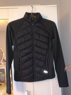 Svart vindjacka med quiltade paneler - Snygg svart vindjacka med quiltade paneler framtill och släta ärmar från Lager 157. Jackan har hel dragkedja, hög krage och reflekterande logga på ena sidan. Perfekt för blåsiga dagar och sportiga outfits. Har använts några gånger men ser ut som den är helt ny! 