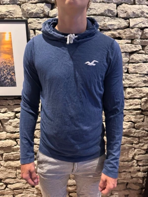 Blå hoodie från Hollister - Mysig blå hoodie från Hollister med vit logga på bröstet och dragsko i huvan. Tillverkad i ett mjukt och tunt material som passar perfekt för en avslappnad stil. Långärmad modell med enkel design och skön passform.
