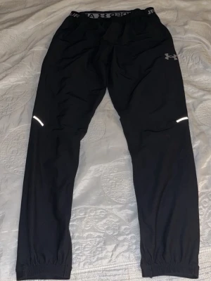 Svarta träningsbyxor Under Armour - Säljer ett par svarta träningsbyxor från Under Armour med resår i midjan och logga på vänster lår. Byxorna har smal passform, reflexdetaljer på benen och elastiska benslut. Perfekta för löpning eller gym. Materialet är lätt och andas.