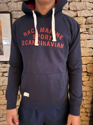 Mörkblå hoodie Race Marine - Mörkblå hoodie från Race Marine med röd text 'RACE MARINE SPORT SCANDINAVIAN' på bröstet. Klassisk känguruficka framtill, vita dragsnören och huva med röd insida. Mjuk bomullsblandning och ribbade muddar vid ärmar och nederkant.