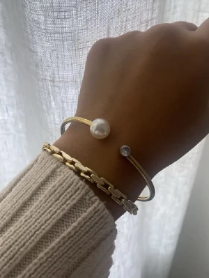 Guldfärgat armband med pärla - Stilrent guldfärgat armband i metall med öppen design. Ena änden pryds av en vit pärla och den andra av en klar sten. Perfekt för dig som gillar minimalistiska smycken med en elegant touch.