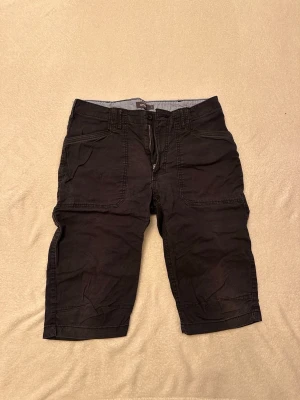 Knåläng cargoshorts med fickor - Säljer ett par svarta cargoshorts under knäna från dressman med flera praktiska fickor både fram och bak. Shortsens material känns slitstarkt och de har bälteshällor i midjan. Perfekta för dig som gillar en avslappnad och funktionell stil.