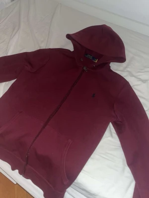 Vinröd hoodie från Polo Ralph Lauren - Snygg vinröd hoodie med dragkedja från Polo Ralph Lauren. Klassisk design med huva, känguruficka och broderad logga på bröstet. Perfekt för en avslappnad och stilren look.