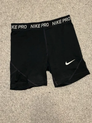 Svarta Nike Pro träningsshorts - Säljer ett par svarta Nike Pro shorts med elastisk midja där det står 'NIKE PRO' runtom. Shortsen har en tight passform och är perfekta för träning. Klassisk vit Nike-logga på ena benet och sömmar som ger en sportig look.
