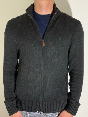 Mörkgrå ziptröja från Ralph Lauren - Mörkgrå stickad ziptröja från Ralph Lauren med hög krage och diskret broderad logga på bröstet. Tröjan har ribbade muddar och nederkant samt dragkedja framtill. Perfekt för lager-på-lager och snygg till jeans.
