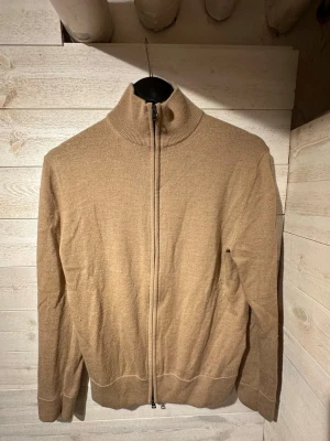 merinoullströja från these glory days - Snygg beige tröja från These Glory Days i mjuk superior merinoull. Tröjan har hög krage, hel dragkedja framtill och ribbade muddar vid ärmslut och nederkant. Perfekt för dig som gillar stilrena och bekväma plagg.