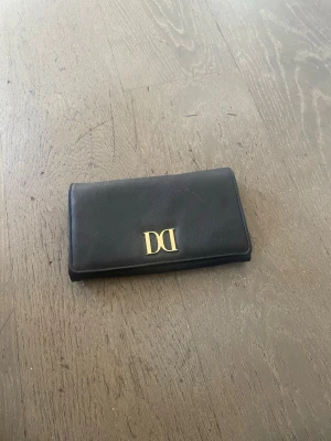 Svart clutch med gulddetalj - Snygg svart clutch i läderimitation med guldfärgad DD-logga på framsidan. Plånboken har en klassisk rektangulär form och stilren design som passar perfekt i väskan. Enkel att matcha med olika outfits och praktisk för kort och kontanter.