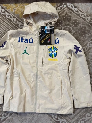 Brasil windbreaker Jordan storlek S - Stilren beige vindjacka från Jordan med Brasil CBF-logga, blåa och gula detaljer samt sponsormärken. Jackan har huva, hel dragkedja och är tillverkad i slitstarkt Gore-Tex-material som står emot vind och regn. Perfekt för dig som gillar sportig streetwear och fotbollsinspirerad stil. Passar som storlek S