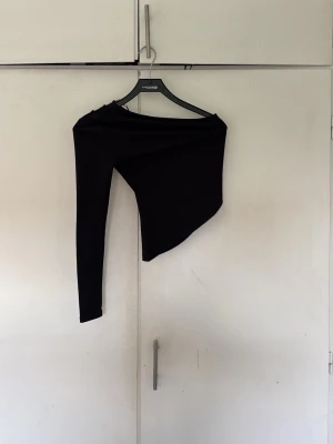 Svart oneshoulder topp - Snygg svart oneshoulder topp med lång ärm på ena sidan och axelbandslös på andra. Perfekt för dig som vill ha en unik och trendig look. Tunn och stretchig modell som sitter tight mot kroppen.