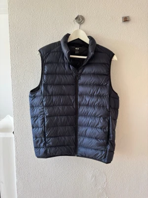 Mörkblå dunväst från Uniqlo - Snygg mörkblå dunväst från Uniqlo med hög krage och dragkedja framtill. Västen har en quiltad design med horisontella sömmar och är lätt vadderad för extra värme. Perfekt att slänga över en hoodie eller tröja för en chill look.