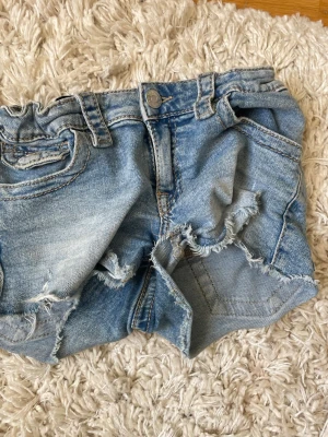 Ljusa blå jeansshorts med slitningar - Säljer ett par ljusa blå jeansshorts med råa, slitna detaljer och fransiga kanter. Shortsen har klassisk femficksdesign, knappgylf och bälteshällor. Perfekta för sommaren och ger en avslappnad look.