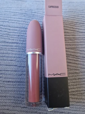 MAC Capricious flytande läppstift - MAC Capricious är ett flytande läppstift i en snygg, dammigt rosa nyans. Förpackningen har en matt rosa kork och genomskinlig kropp som visar färgen. Märket syns tydligt på både hylsa och kartong. Perfekt för dig som gillar en mjuk, naturlig look.