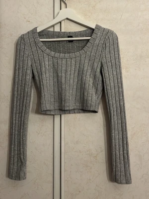 Grå ribbad croppad långärmad topp - Grå croppad topp med långa ärmar och bred ribbad struktur. Toppen har en rund halsringning och är tillverkad i ett mjukt, stretchigt material som sitter snyggt på kroppen. Perfekt att styla med höga jeans eller kjol.