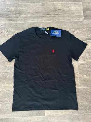 Svart t-shirt från Polo Ralph Lauren - Klassisk svart t-shirt från Polo Ralph Lauren med rund hals och korta ärmar. T-shirten har den ikoniska röda logotypen broderad på bröstet och är tillverkad i mjuk bomull för en skön känsla. Perfekt till jeans eller shorts. Storlek M 