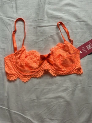 Orange spets-bh - Säljer en snygg orange bh, i genomskinlig spets med dekorativ rosett framtill. Bh:n har justerbara axelband och bygel för extra stöd. Perfekt för dig som gillar färgglada och trendiga underkläder med fina spetsdetaljer.