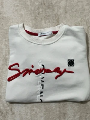 Vit Givenchy sweatshirt med röd text - Säljer en vit sweatshirt från Givenchy med röd broderad text och vertikal logotyp i vitt på framsidan. Tröjan har rund halsringning och långa ärmar. Materialet är mjukt och bekvämt, perfekt för en trendig och avslappnad stil. Skickar samma dag vid köp.