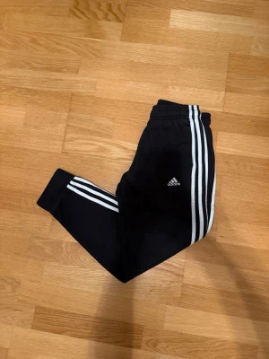 Svarta Adidas mjukisbyxor med vita ränder storlek 152  - Säljer ett par svarta mjukisbyxor från Adidas med de klassiska tre vita ränderna längs benen och Adidas-logga på låret. Byxorna har resår i midjan och vid bensluten för en skön passform. Perfekta för chill eller träning.