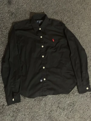 Svart skjorta från Ralph Lauren xs - Klassisk svart skjorta från Ralph Lauren med vit knappstängning och röd broderad logga på bröstet. Skjortan har lång ärm och normal passform, perfekt till en clean och stilren look det står att det är XL men passar som xs pris kan diskuteras vid snabb affär .