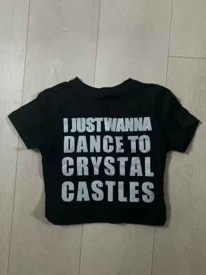 Svart croppad t-shirt baby tee Crystal Castles - Svart croppad t-shirt med vit text 'I JUST WANNA DANCE TO CRYSTAL CASTLES' på framsidan. T-shirten har korta ärmar och rund hals. Perfekt för dig som gillar bandmerch och vill sticka ut med ett statement-plagg.