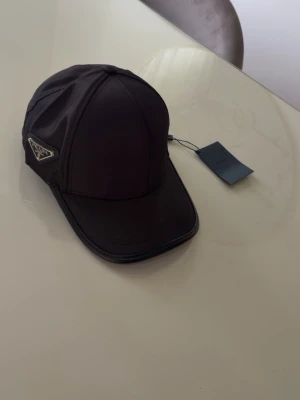 Svart keps från Prada i nylon - Snygg svart keps från Prada i glansigt nylonmaterial med klassisk böjd skärm och Pradas ikoniska metallogo på sidan. Kepsen har sex paneler och justerbar rem bak. Perfekt accessoar för en trendig och stilren look.