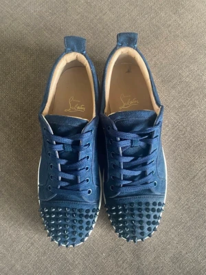 Blå sneakers från Louboutin - Snygga blå sneakers från Christian Louboutin i mocka med coola svarta nitar på tån. Skorna har vita sulor, blå skosnören och en låg siluett. Insidan är beige och märkt med Louboutin-loggan. Endast skorna följer med, därav priset.