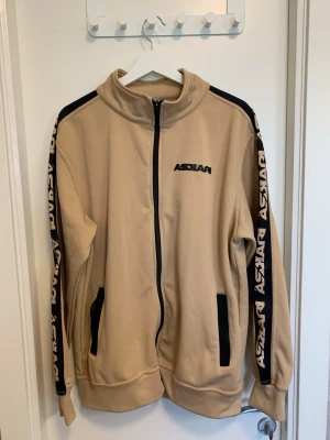 Askari tracksuit Naz Black - Snygg beige track jacket från Askari med svarta detaljer och logga på bröstet samt längs ärmarna. Jackan har hel dragkedja framtill, två fickor och hög krage. Tillverkad i mjukt syntetmaterial och perfekt för träning eller chill. Både tröjan och byxorna. Använd 2 gånger. Skambud undanbes