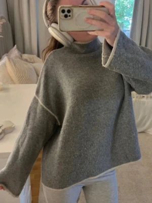 Grå stickad tröja med vit kant - Mysig grå stickad tröja med hög krage och vita kontrastsömmar längs ärmar och kanter. Tröjan har en avslappnad passform och långa ärmar, perfekt för kyliga dagar. Enkel och stilren design som passar till många outfits💕🌹