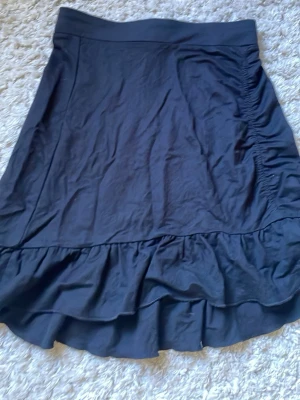 Svart volangkjol från Gina Tricot - Säljer en svart kjol från Gina Tricot med bred midja och volangkant nedtill. Kjolen har en enkel, stilren design med lätt rynkade detaljer på sidan och är perfekt för dig som gillar en klassisk look med lite extra volym.