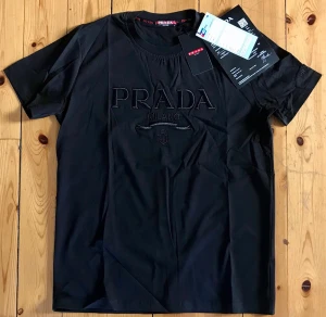 Svart t-shirt från Prada Milano - Snygg svart t-shirt från Prada Milano med broderad logga framtill. Klassisk rund halsringning och korta ärmar. Tillverkad i mjuk bomull för en skön känsla. Perfekt för dig som vill ha en stilren och exklusiv look.