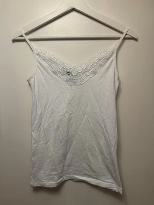 Vit spetsdetaljerad linnetopp Vero Moda - Säljer en vit linnetopp från Vero Moda med tunna axelband och vacker spets längs urringningen. Toppen är ärmlös och har en feminin, enkel design som passar perfekt till sommaren. Materialet är mjukt och bekvämt.