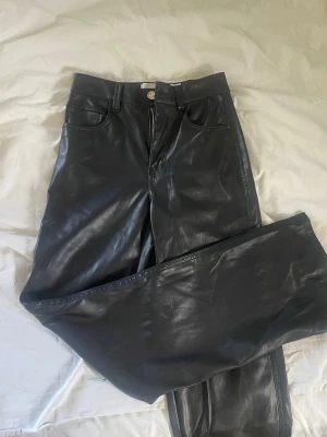 Svarta fuskskinnbyxor med raka ben - Snygga svarta byxor i fuskskinn med raka ben och klassisk femficksmodell. Byxorna har hög midja, bälteshällor och dragkedja med knapp framtill. Perfekta för dig som vill ha en edgy look och gillar trendiga material.