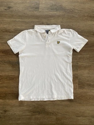 Lyle & Scott piké - stl S - Klassisk vit pikétröja från Lyle & Scott med korta ärmar och krage. Tröjan har knäppning framtill och den ikoniska gula loggan broderad på bröstet. Perfekt för en stilren och avslappnad look.