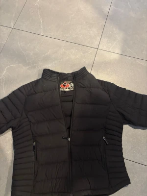 Svart pufferjacka från Moose Knuckles - Säljer en svart pufferjacka från Moose Knuckles med coolt camo-märke på insidan. Jackan har ståkrage, två dragkedjeförsedda fickor och quiltad design som ger en schysst look. Perfekt för kalla dagar och riktigt snygg till streetwear-stilen.
