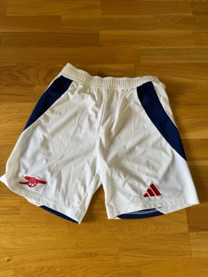 Vita Arsenal shorts från Adidas - Säljer ett par vita Arsenal shorts från Adidas med marinblå detaljer på sidorna och röda loggor. Shortsen har elastisk midja och är tillverkade i ett lätt och svalt syntetmaterial, perfekta för fotboll eller träning. Passar som S. 