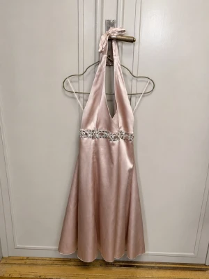 Champagnefärgad halterneckklänning - Snygg champagnefärgad klänning i glansigt satintyg med halterneck och djup urringning. Midjan är dekorerad med stora glittriga stenar som ger extra bling. Storlek 14. Den har en fläck på insidan