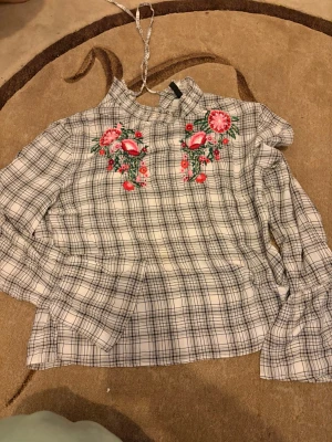 Rutig blus med broderi från H&M - Supersnygg rutig blus från H&M med hög krage och långa ärmar. Blusen har broderade blommor i rosa och rött framtill. Materialet är lätt och skönt, perfekt till jeans eller kjol. Strl 38