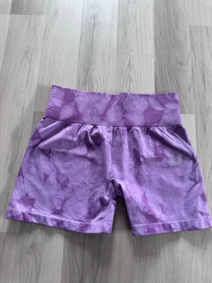 Lila träningstights-shorts från ICANIWILL - Säljer ett par lila träningstights-shorts från ICANIWILL med bred ribbad midja och snyggt batikmönster. Shortsen har en högmidjad passform och är tillverkade i ett stretchigt syntetmaterial. Använda som max 2 gångener. 