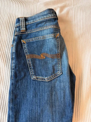 Blå jeans från Nudie Jeans Co - Snygga blå jeans från Nudie Jeans Co med klassisk femficksmodell och orange kontrastsömmar. Bakfickan har den ikoniska vågiga sömmen. Jeansen är i ett slitstarkt denimtyg och har knappgylf. Perfekta för en avslappnad och trendig look. W 26 L 32