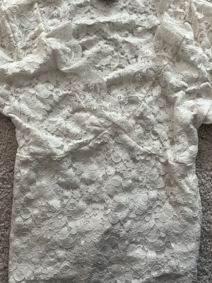 Vit spetsblus med v-ringning - Säljer en vit spetsblus från GINA TRICOT med v-ringning och korta ärmar. Blusen har ett romantiskt blommigt spetsmönster och är lätt transparent. Perfekt att styla med jeans eller kjol för en trendig look.