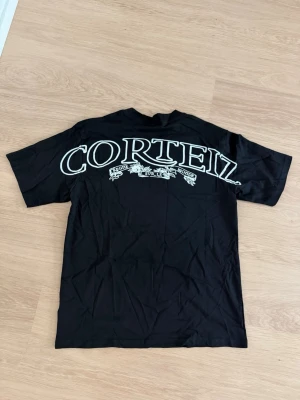 Svart Corteiz t-shirt med tryck - Svart t-shirt från Corteiz med stort vitt tryck på ryggen och mindre logga framtill. Klassisk passform med rund hals och korta ärmar. Perfekt för dig som gillar streetwear och vill sticka ut med en snygg logga.