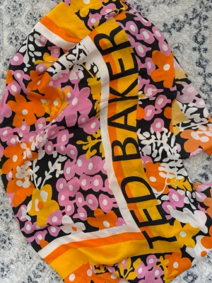 Blommig scarf från Ted Baker - Färgstark scarf från Ted Baker med stora blommor i rosa, orange, gult och vitt på svart botten. Tydlig logga längs kanten och kontrasterande ränder i orange och vitt. Tunn och mjuk, perfekt att styla som topp eller runt halsen.