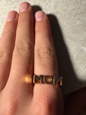 Guldfärgad ring med MOM-text - Unik ring i metall med guldfärgad finish och texten 'MOM' i stora bokstäver framtill.men är tyvärr lite sliten.